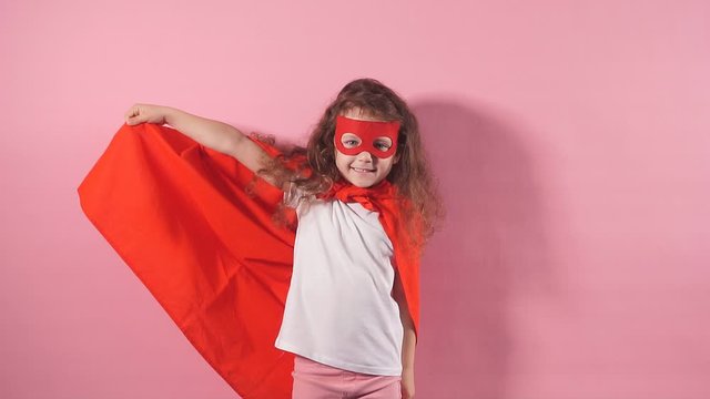 Portrait of confident smiling kid superhero wearing red cloak and mask on eyes imagines herself as hero, stand isolated on pink background