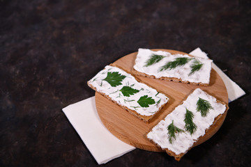 Crispbread with curd cheese and herbs, on a dark background, on a wooden board. Space for text....