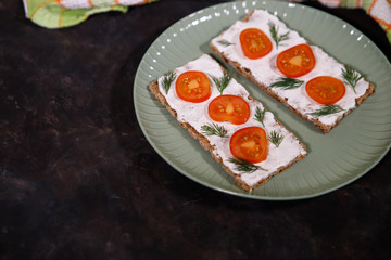 Crispbread with curd cheese and cherry tomato lies on a plate, on a dark background. Space for text. Flat lay, top view. Diet snack.