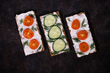 Crispbread with curd cheese, cherry tomato and cucumber, on a dark background. Space for text. Flat lay, top view. Diet snack.