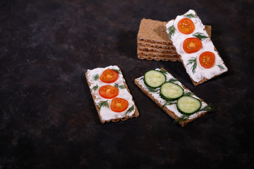 Crispbread with curd cheese, cherry tomato and cucumber, on a dark background. Space for text. Flat lay, top view. Diet snack.