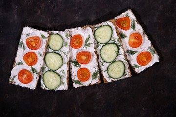 A set of crispbread with curd cheese and fresh vegetables, cherry tomato and cucumber, on a dark background. Space for text. Flat lay, top view. Diet snack.