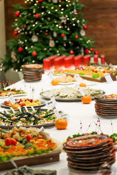 Christmas dishes and snacks at the party. Blurred decorated fir tree on the background. Bright festive food picture. Red drinks in glasses, sturgeon, red caviar, tangerines. Disposable plates