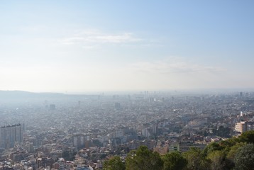 Fototapeta na wymiar Vista aérea de la ciudad de Barcelona con luz natural de día, y con un cielo despejado de fondo y distintos árboles enmarcando la parte inferior de la imágen
