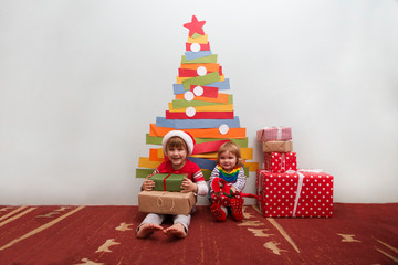 Obraz na płótnie Canvas Two children brother and sister in Santa red hat with gifts near homemade Christmas tree made of paper on wall. Christmas time. Eco frendly decor for Christmas. Zero waste Christmas