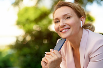 Smiling woman with smartphone on the street.  Happy businesswoman is using phone, outdoors. Cheerful businesswoman in a jacket with cell phone in park.