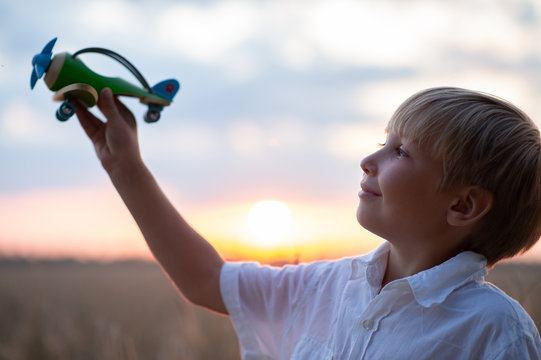 Happy child playing with a toy plane in nature during summer sunset. Boy in a  white shirt with a plane in hands on wheat field. Kid holds a wooden airplane and dreams of being a pilot, on the nature - Powered by Adobe
