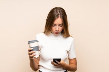 Young blonde woman over isolated background holding coffee to take away and a mobile