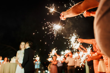 A crowd of young happy people with sparklers in their hands during celebration. Sparkler in hands on a wedding - bride, groom and guests holding lights in hand. Sparkling lights of bengal fires.