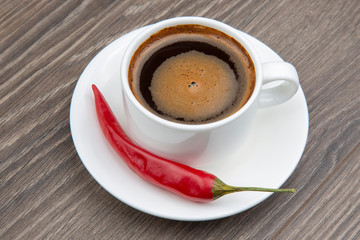 Black coffee in a white cup with red hot pepper on a plate. Spicy food and strong drink.