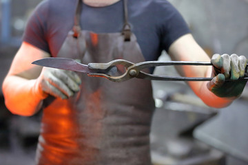 Worker in the blacksmith shop shows tongs with forged preheated workpiece