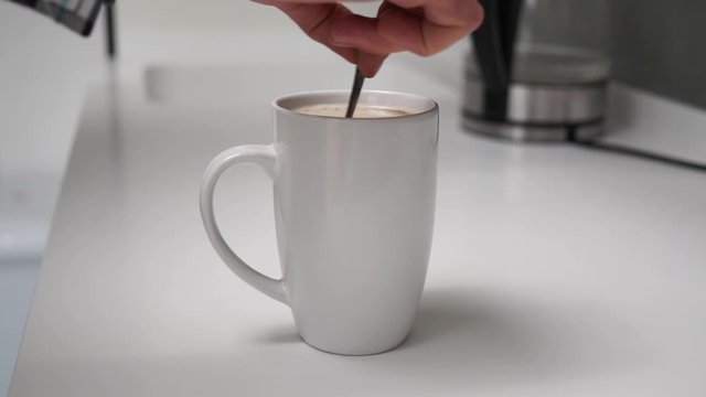 Side view of person hand stiring sugar in white cup of black coffee with milk with silver spoon on white table in a modern white high-tech kitchen or cafe. Cappuccino or tea in the mug
