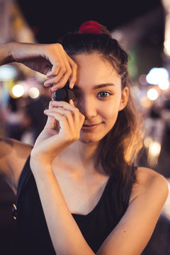 Fototapeta Girl holding small black cemara look pretty to pretend to take a shot, one big eye with smiling face, outdoot at night have some bokeh of light on background