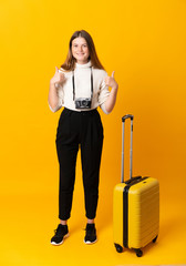 Full body of traveler teenager girl with suitcase over isolated yellow background giving a thumbs up gesture
