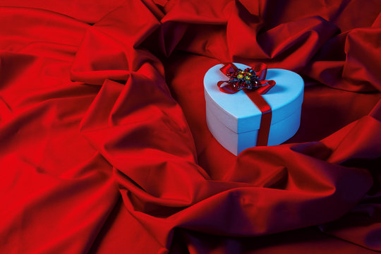 valentines greeting card in blue light. white cardboard box in shape of heart wrapped in ribbon lay on a red cloth which repeats the form of present package. love and romance gift concept