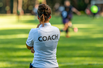 Back view of a female sport coach watching her team compete at an outdoor football field - obrazy, fototapety, plakaty