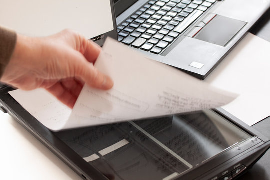 A Hand Puts A Piece Of Paper On A Flatbed Scanner With A Laptop Computer In The Background. Selective Focus And Hand In Motion Blur.