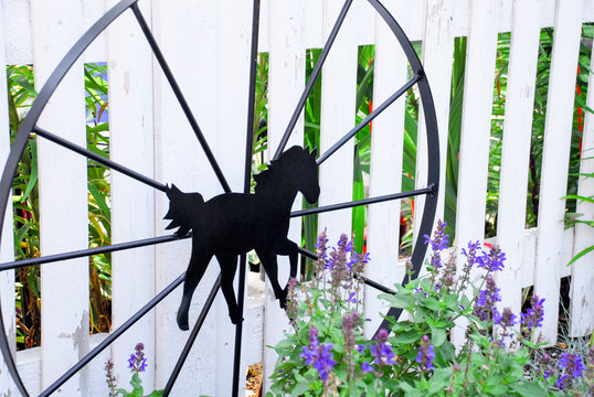 Garden ornament of black iron wagon wheel with black horse in middle hanging on white picket fence with garden flowers