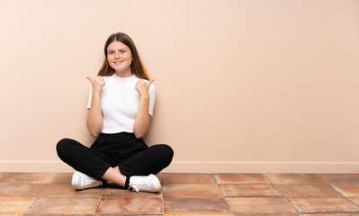 Ukrainian teenager girl sitting on the floor with thumbs up gesture and smiling