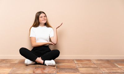 Ukrainian teenager girl sitting on the floor extending hands to the side for inviting to come