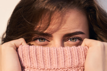 A close-up of green eyes of woman who has lifted pink knitted sweater to her nose, holding on to its edges so that only look is visible. Natural beauty, clear portrait of girl, gaze.