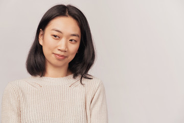 Close-up face, slightly turned to the side, a thoughtful look directed down. Businesslike young woman Asian appearance dressed in knitted warm sweater stands isolated white background in Studio.