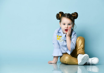 Surprised little baby kid girl in blue shirt and gold leather pants is sitting on the floor holding hand at her cheek