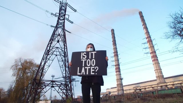 Is It Too Late? Beautiful girl standing near working factory holding poster in face protection mask looking at camera worried face. Nature protection. Global warming potential.Air pollution prevention