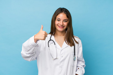 Young brunette girl over isolated blue background with doctor gown and with thumb up