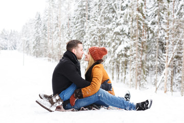 Fototapeta na wymiar Couple in love, sitting on the snow-covered ground, hugging, looking at each other, a background of snow-covered trees and firs, love story