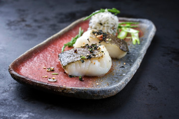 Gourmet fried Japanese skrei cod fish filet with glasswort and furikake onigiri as closeup on a modern design plate