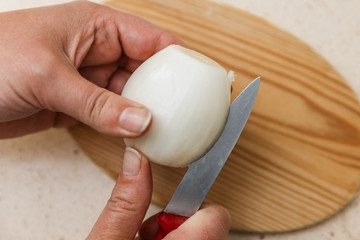 dos manos pelan una cebolla con un cuchillo encima de una tabla de cortar de madera, encima de mesa de mármol de la cocina.