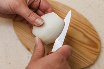 dos manos pelan una cebolla con un cuchillo encima de una tabla de cortar de madera, encima de mesa de mármol de la cocina.