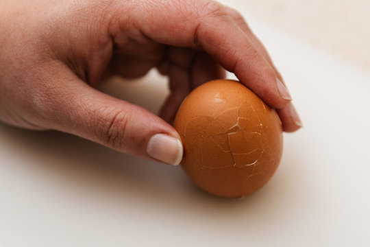 una mano de mujer esta rompiendo la c&aacute;scara de un huevo duro de gallina sobre tabla de corte blanca