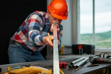A young guy engineer in a plaid shirt and an orange helmet checks the straightness of the metal profile by eye.