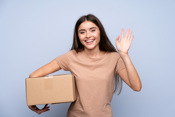 Young woman over isolated blue background holding a box to move it to another site and saluting