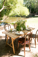 Festive table decor in openair next to the pond. Green color with wooden cutlery. Different natural white flowers. Wooden table and chairs. Wine glasses. Luxury wedding, party, birthday.