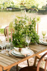 Festive table decor in openair next to the pond. Green color with wooden cutlery. Different natural white flowers. Wooden table and chairs. Wine glasses. Luxury wedding, party, birthday.