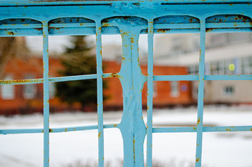 A metal fence lattice covered with cracked blue paint and rust, behind the fence, in the background lies snow and blurry buildings.
