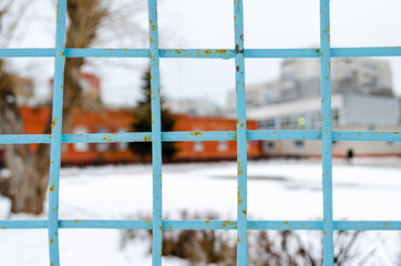 A metal fence lattice covered with cracked blue paint and rust, behind the fence, in the background lies snow and blurry buildings.