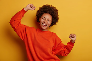 Photo of joyful brunette woman has fun and dances with hands raised, dressed in casual jumper,...