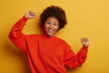 Upbeat dark skinned young woman raises both arms, feels lucky, attends awesome party, dressed in oversized jumper, stands against yellow studio wall. Lifestyle, celebration, wellbeing concept