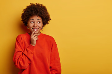 Fototapeta na wymiar Charming tender curly woman smiles gladfully, looks aside, wears casual clothes, stands against yellow studio wall, free space for your advertising, satisfied with good promo, being amused and lucky