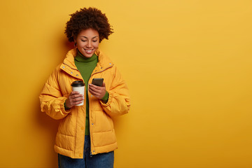 Photo of attractive positive woman in yellow outerwear, happy to read good comments under post, holds modern cellphone, drinks takeout coffee, checks newsfeed during break. People and lifestyle