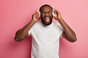 Fototapeta na wymiar Emotional relaxed carefree bearded man listens favourite song from playlist, entertains indoor, wears white headphones on ears, casual t shirt, poses over pink wall. Happy male meloman enjoys music