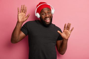 Satisfied bearded male youngster listens merry song in headphones, moves over pink background, boosts mood with cool music, feels upbeat, wears red hat and black t shirt. Having fun during free time