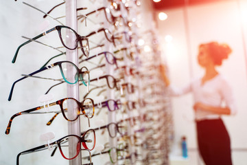 Eyeglasses shop. Stand with glasses in the store of optics. Woman on blurred background chooses glasses. closeup - obrazy, fototapety, plakaty