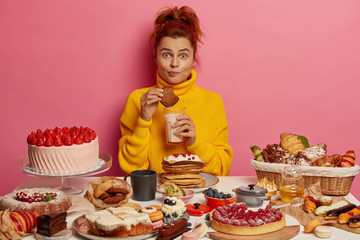 People, nutirtion, calories, bakery concept. Ginger girl in yellow jumper eats tasty oatmeal cookies and drink yoghurt, sits at table with many delicious cakes, doesnt keep to diet. Sugar addiction