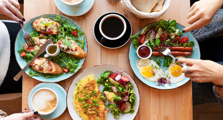 Different colorful meals for breakfast or lunch time on a plate with cutlery on woman's hands. Fried eggs, omelette, bruschetta and sausage on a wooden teble in restaurant. Flat lay top view panoramic