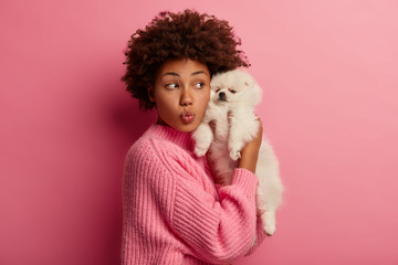 Lovely ethnic woman holds white miniature puppy closely to face, keeps lips rounded, dressed in knitted sweater, expreses love to and care to favourite pet, isolated over pink wall. Animals concept